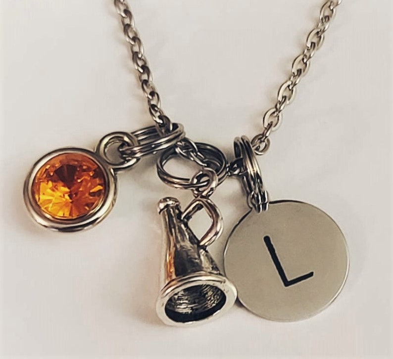 May include: Silver-toned necklace with multiple charms. Charms include a round orange gemstone, a megaphone, and a circular pendant with the letter "L". The necklace is on a white background.