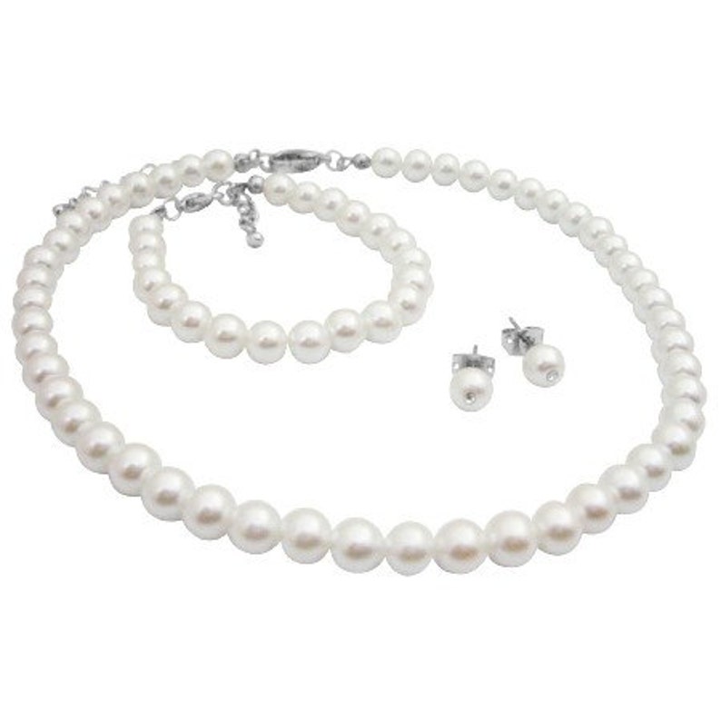 Puede incluir: Un conjunto de joyas de perlas blancas que incluye un collar, una pulsera y pendientes de bot&oacute;n. El collar y la pulsera presentan una hebra de perlas blancas redondas. Los pendientes tienen una sola perla en un poste plateado. El conjunto est&aacute; sobre fondo blanco.