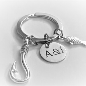 May include: Silver-toned keychain featuring a fishing hook charm, a fish charm, and a round charm engraved with "A & I". The keychain is designed for keys and accessories.