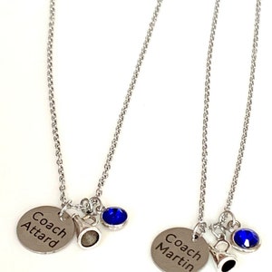 May include: Two silver-toned necklaces with chain links. Each necklace has a round charm engraved with a name, a small megaphone charm, and a blue gemstone. One charm reads "Coach Attard", the other reads "Coach Martin".