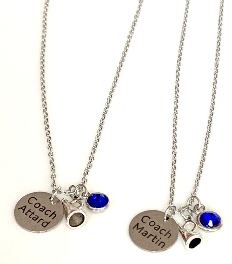May include: Two silver-toned necklaces with chain links. Each necklace has a round charm engraved with a name, a small megaphone charm, and a blue gemstone. One charm reads "Coach Attard", the other reads "Coach Martin".