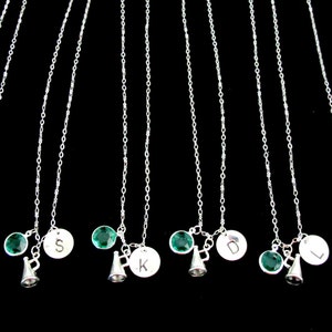 May include: A collection of silver necklaces with personalized initial pendants, green gemstone accents, and small megaphone charms. The necklaces hang against a black background, showcasing the delicate chains and charms.