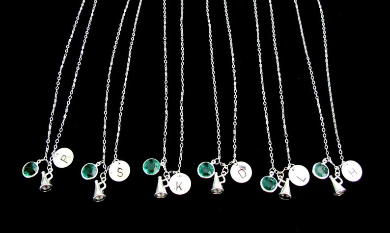 May include: A collection of silver necklaces with personalized initial pendants, green gemstone accents, and small megaphone charms. The necklaces hang against a black background, showcasing the delicate chains and charms.