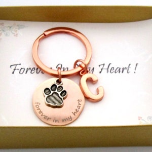 May include: A copper-colored keychain with a paw print charm and the letter "C" is presented in a gold gift box. The charm reads "forever in my heart." The background includes a card with the words "Forever In My Heart!"