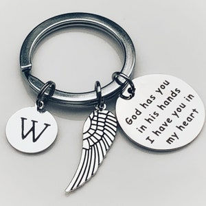 May include: A silver keychain with a large ring holding three charms. One charm is a round disc with the letter "W", another is a wing, and the third is a round disc with the text "God has you in his hands I have you in my heart."