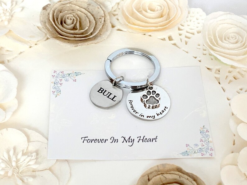 May include: Silver keychain with two round charms. One charm has the name "BULL" engraved, the other has a paw print and the words "forever in my heart". The keychain is on a card that says "Forever In My Heart".