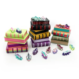 May include: A collection of miniature dragon figurines in various colors and patterns, displayed alongside several stacks of small, colorful, rectangular book-shaped boxes. The dragons feature textured scales and small horns, arranged on and around the books.
