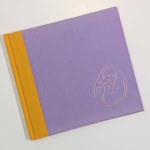 May include: A square hardcover book with a light purple cover and a vertical mustard yellow spine strip. The front cover features a thin, gold-colored line drawing of a fetus centered in the lower right quadrant, enclosed within a circular outline.
