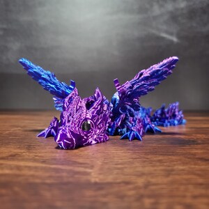 Puede incluir: Una pequeña figura de dragón de color morado y azul con alas detalladas y una superficie texturizada. El dragón tiene un pequeño ojo negro y está posado con las alas extendidas.