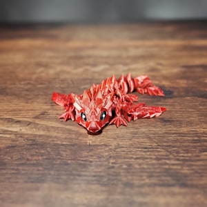 Może przedstawiać: Czerwony, segmentowy, plastikowy smok z kolczastym grzbietem i szerokim, zębatym uśmiechem. Smok leży na drewnianej powierzchni.