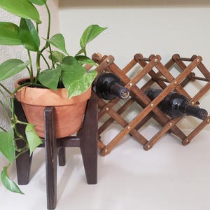 May include: A terracotta pot with a green plant sits on a dark brown wooden plant stand. A wooden wine rack holds two bottles of wine. The wine rack is made of interlocking wooden slats.
