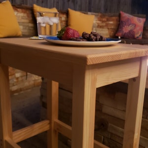 May include: A light wood end table with a simple design. The table has a rectangular top and four legs. The table is sitting on a patio with a brick wall in the background.