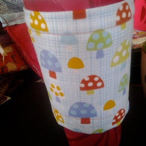 May include: A red disposable coffee cup with a white lid and a fabric sleeve. The sleeve is light blue with a pattern of colorful mushrooms. The mushrooms have red, yellow, blue, and green caps.