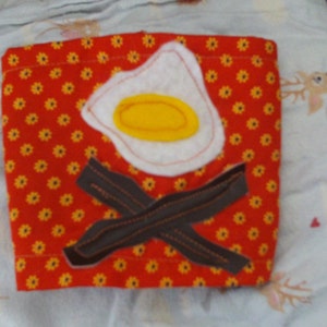 May include: A square, red fabric item with a repeating yellow floral pattern. It features an appliqued fried egg with a yellow yolk and a white outline, and two strips of brown bacon. The item is likely a kitchen accessory.