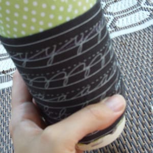 Könnte beinhalten: Ein Pappbecher mit dekorativer Hülle. Die Hülle ist dunkelbraun mit weißer Schreibschrift und gepunkteten Linien. Der obere Teil des Bechers hat ein grünes Tupfenmuster.