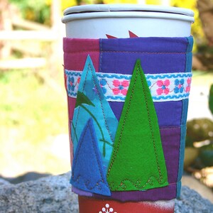 May include: A reusable coffee cup with a colourful, patchwork-style cosy. The cosy features appliquéd felt trees in blue and green, with a floral ribbon accent. The cup is red with white snowflake designs.