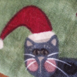 May include: Close-up of a fabric with a repeating pattern of a cat wearing a Santa hat. The cat is dark blue with pink cheeks and white whiskers. The hat is red with a white trim, set against a green background.
