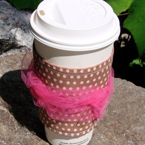 Op de afbeelding: Een witte papieren koffiebeker met een wit deksel. De beker heeft een bruine polka dot mouw en een roze tule wikkel. De tekst op de beker luidt: "Pas op, de drank die je gaat nuttigen is extreem heet."