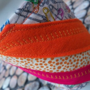 Pode incluir: Close-up de um artigo de tecido feito à mão com camadas de tecido estampado e feltro. A camada superior apresenta uma impressão de alfinetes de segurança, a camada do meio é feltro laranja com detalhes costurados e a camada inferior é uma fita rosa.