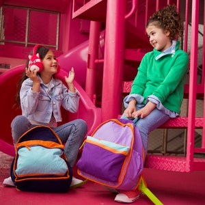 Puede incluir: Dos niños, uno con una chaqueta vaquera azul y el otro con un suéter verde, están sentados en una estructura de juegos rosa. Ambos llevan mochilas, una morada y otra azul con detalles naranjas. El niño con la chaqueta vaquera sostiene un juguete de peluche rojo en forma de zorro con auriculares.