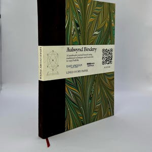 Op de afbeelding: Een handgemaakt notitieboek met een gemarmerde groene, gele en witte kaft en een donkerbruine rug. Het notitieboek heeft een rood lint als bladwijzer en een witte band met de tekst "Aulwynd Bindery" en "Lined Ivory Paper".