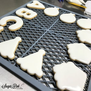 May include: White frosted sugar cookies in various shapes, including letters, ornaments, and party hats, are arranged on a gray cooling rack. The cookies have a light brown base and are decorated with a smooth, glossy white frosting. The logo "Sugar Dot Cookies" is visible in the bottom left corner.