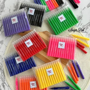 May include: A collection of colorful edible ink markers in various shades, including purple, black, green, pink, red, blue, orange, and yellow. Each set is packaged in clear plastic and features a label with the text "Sugar Dot" and "Edible Ink Marker".