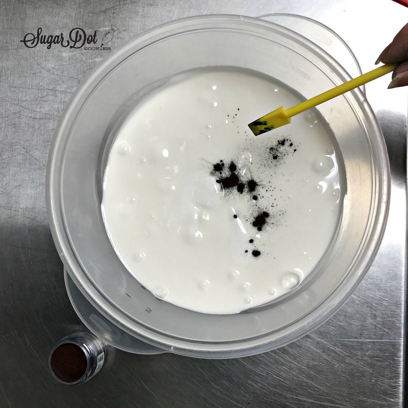 Peut inclure: Un bol en plastique transparent rempli de liquide blanc, peut-&ecirc;tre du gla&ccedil;age, avec du colorant alimentaire brun fonc&eacute; ajout&eacute;. Une spatule jaune est utilis&eacute;e pour m&eacute;langer les ingr&eacute;dients. Le logo Sugar Dot Cookies est dans le coin sup&eacute;rieur gauche.