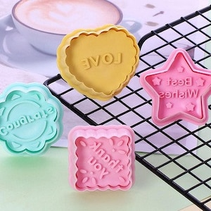 May include: Four colorful cookie cutters with different shapes and messages. A green flower-shaped cutter says "Celebrate", a pink square cutter says "Thank You", a yellow heart-shaped cutter says "Love", and a pink star-shaped cutter says "Best Wishes".