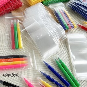 May include: Assortment of clear plastic zip-top bags and colorful edible ink markers. Markers are in various colors, including red, green, yellow, orange, pink, and blue. The text "Sugar Dot" and "Edible Ink Marker" are visible on the markers.