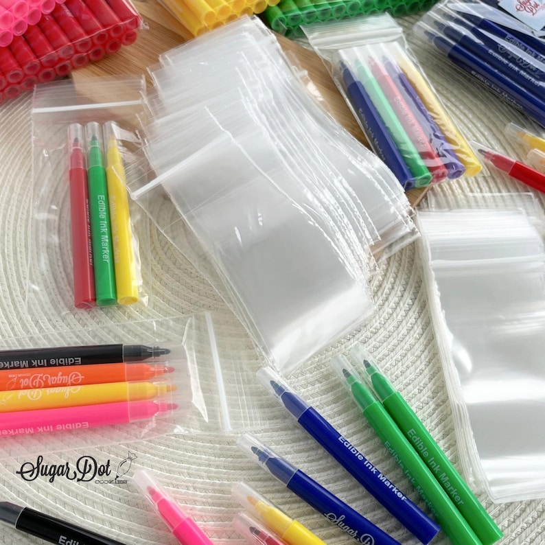 May include: Assortment of clear plastic zip-top bags and colorful edible ink markers. Markers are in various colors, including red, green, yellow, orange, pink, and blue. The text "Sugar Dot" and "Edible Ink Marker" are visible on the markers.