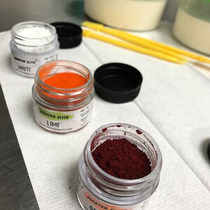 Peut inclure: Plusieurs pots en verre de colorants alimentaires Master Elite. Les couleurs comprennent le blanc, l'orange et un rouge-orange fonc&eacute;. Les pots sont ouverts, r&eacute;v&eacute;lant les pigments vibrants. Les textes "LIME" et "ORANGE" sont visibles sur les &eacute;tiquettes.