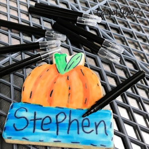 May include: A decorated pumpkin cookie with the name "Stephen" written on a blue rectangle. Several black edible ink pens with clear caps are scattered around the cookie on a gray grid.