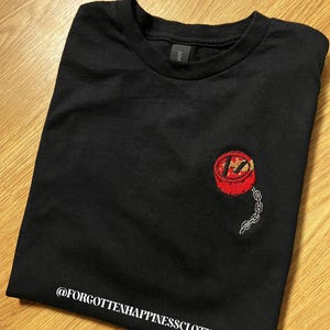 Puede incluir: Camiseta negra con un diseño bordado en rojo y dorado con una cadena. El texto "@FORGOTTENHAPPINESSCLOTHING" está impreso en blanco en la parte inferior. La camiseta está doblada sobre una superficie de madera.