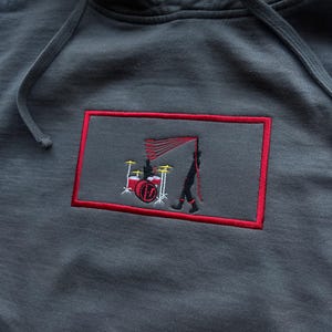 May include: Dark grey hoodie with a red-bordered rectangular patch. The patch features an embroidered design of a drummer and a person holding a flag, in red and black. The hoodie has a drawstring hood.