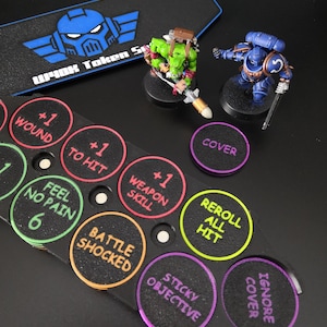 Peut inclure: Un support de jetons de jeu en plastique noir avec des emplacements circulaires contenant divers jetons colorés marqués de texte, notamment +1 WOUND, +1 TO HIT, +1 WEAPON SKILL, FEEL NO PAIN 6, BATTLE SHOCKED, REROLL ALL HIT, STICKY OBJECTIVE et COVER IGNORE.
