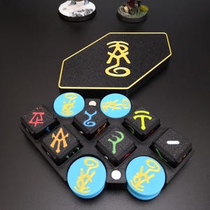 Può includere: Un tabellone di gioco nero con un coperchio bordato d'oro e un vassoio contenente dadi e gettoni. I dadi sono neri con simboli colorati e i gettoni sono blu con disegni dorati. Due miniature sono sullo sfondo.