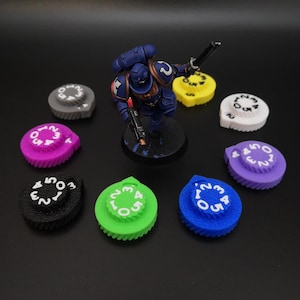 Op de afbeelding: Een verzameling kleurrijke, genummerde wijzerplaten in verschillende kleuren, waaronder grijs, roze, zwart, groen, blauw, paars, geel en wit. Een blauw miniatuurfiguur staat in het midden. De wijzerplaten zijn waarschijnlijk voor een tabletopspel.