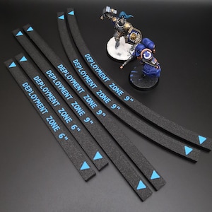 Peut inclure: Marqueurs de zone de déploiement en mousse noire avec texte bleu et indicateurs de flèches. Le texte indique "DEPLOYMENT ZONE 6" et "DEPLOYMENT ZONE 9". Deux figurines miniatures sont également dans l'image.