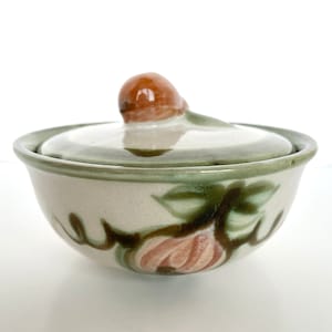 May include: A white ceramic bowl with a lid. The bowl has a green and brown floral design with a pink flower. The lid has a brown knob.