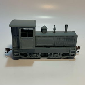 May include: A gray model train locomotive featuring a cab with windows, a central boiler section with a dome, a chimney, and a small vertical detail. The engine has a rectangular body with side paneling, visible wheels, and small couplers at each end.