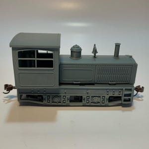 May include: A gray model train locomotive featuring a boxy cab, a central boiler section with a chimney, a small dome, and a whistle. The chassis displays detailed wheels and side panels, with small brown couplers extending from the front and back.
