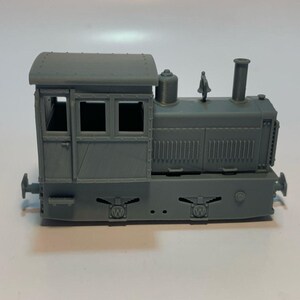 Peut inclure: Une locomotive miniature grise comprenant une cabine détaillée avec des fenêtres, une cheminée verticale, une petite cloche sur le dessus et des panneaux latéraux à lattes verticales. La base présente deux emblèmes circulaires avec un W.