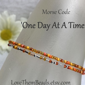 May include: A beaded bracelet with orange, yellow, and silver beads. The bracelet is on a mannequin's wrist. The text "Morse Code 'One Day At A Time'" is above the bracelet. The text "LoveThemBeads.etsy.com" is below the bracelet.