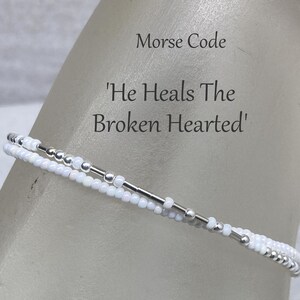 May include: White beaded bracelet with silver beads spelling out the Morse code for 'He Heals The Broken Hearted'.