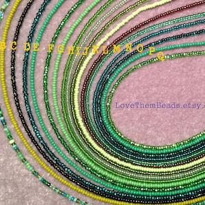May include: A collection of 17 green, blue, and yellow beaded necklaces, arranged in a semi-circle on a pink surface. The necklaces are labeled with letters from A to Q.