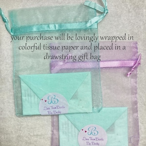 May include: A light blue and purple drawstring gift bag with two light blue envelopes inside. The envelopes have a white circle with the text "Love Them Beads By Becky" in black text. The text "LoveThemBeads.etsy.com" is printed at the bottom of the image.