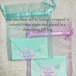 May include: A light blue and purple drawstring gift bag with two light blue envelopes inside. The envelopes have a white circle with the text "Love Them Beads By Becky" and a small heart design.
