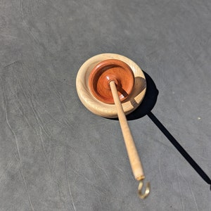 May include: A wooden spinning wheel with a brown wooden base and a light brown spindle with a gold hook. The wheel is on a gray surface.