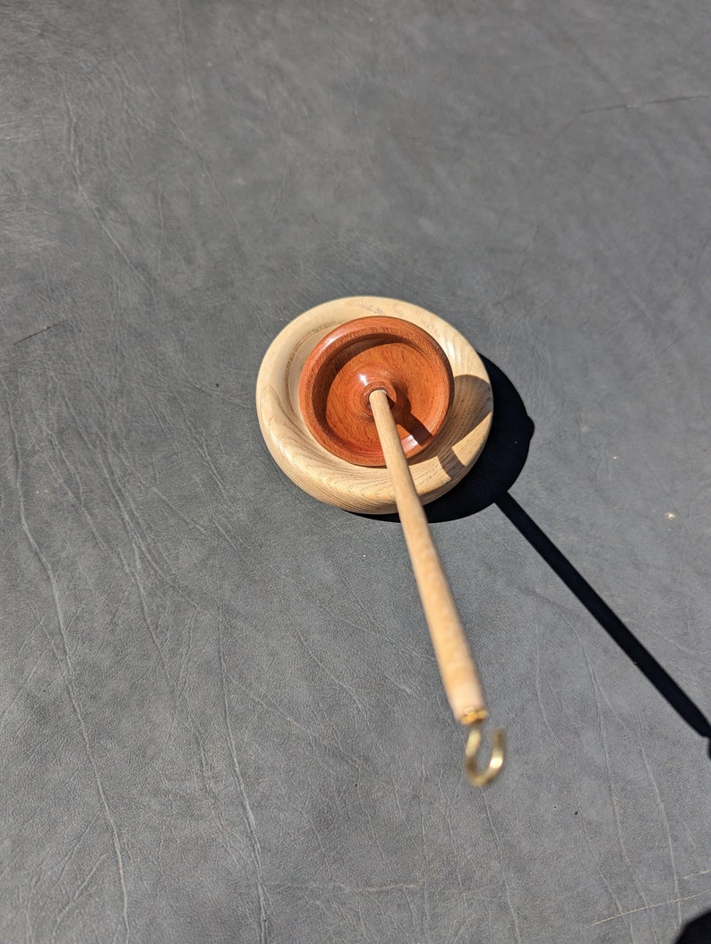 May include: A wooden spinning wheel with a brown wooden base and a light brown spindle with a gold hook. The wheel is on a gray surface.